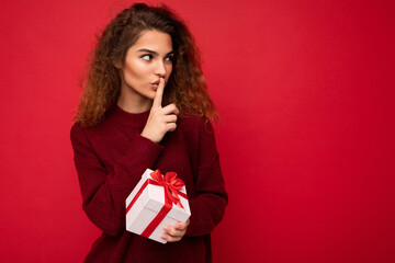 Photo shot of beautiful positive young brunette curly woman isolated over red background wall wearing red sweater holding gift box looking to the side and showing shh gesture