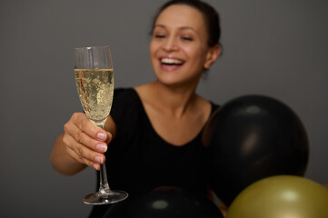 Focus on a sparkling wine in glass in the hand of a blurred woman smiling toothy smile holding gold black air balloons gray background with copy space for advertisement. Celebrating Black Friday
