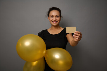 Beautiful smiling happy woman dressed in black outfit shows inflated air balloons of gold color and gold empty blank credit card. Black Friday concept with copy ad space on gray wall background