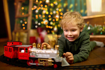Cute curly-haired boy is playing with a magic toy train at the Christmas tree. Concept of...