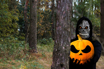 Grim reaper holding halloween pumpkin head. Man in death mask with fire flame in eyes on nature...