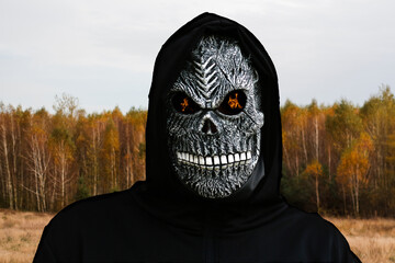 Close-up portrait of grim reaper. Man in death mask with fire flame in eyes on yellow autumn nature sunny background. Carnival costume, creepy teeth. Halloween holiday concept. Dark horror