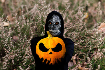 Grim reaper holding halloween pumpkin head. Man in death mask with fire flame in eyes on nature...