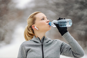 Sportswoman taking a break and drinking water while standing in nature at snowy winter day. Healthy...