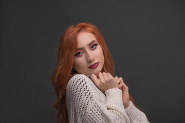 Young beautiful woman with red hair smiles coquettishly from the front with a white sweater on a gray background. Red-haired young woman, smiles with autumn colors.