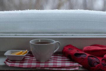cup of tea on the windowsill, slices of lemon lie, a red scarf and gloves are nearby. the window outside is covered with snow, there is a snowdrift.