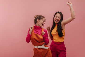 Studio portrait of young asian and european woman happily singing and dancing in pink studio. Slim...