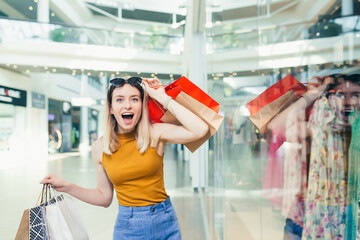 cheerful young shopaholic woman holding paper bags with purchases and smiling looking at camera dancing on shopping center. Happy lady having fun on black friday joyfully jumps for joy in the mall