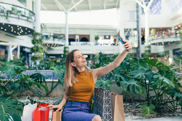 Young woman consumer in the mall browses chat and uses using a smartphone. female standing with a mobile phone in her hands in shopping center. indoor. happy shopper girl with gift bags make purchases