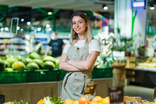 Portrait young woman worker in a Vegetable section supermarket standing smiling with arms crossed. Friendly pleasant female looking at camera in a fruit shop market. Employee in a work apron