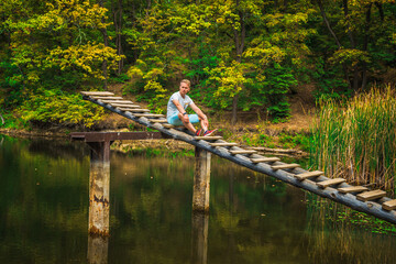 Fototapeta na wymiar A young man on a wooden ladder bridge on a forest mountain lake. Amazing summer landscape with a lake and a panoramic view of a mountain lake
