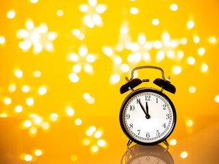 Black clock showing midnight on yellow background with star shaped bright bokeh lights. Time for New year wishes and rituals concept with copy space. New year coming in five minutes countdown mock up.