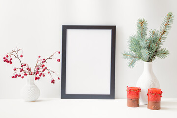 Christmas home interior with decor elements. Mockup with a black frame, branches with red berries in a glass vase, candle on a light background