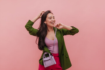 Studio filmed cuddly young asian brunette girl standing against pink background. Model with good white skin smiles as she touches her chin with fingers and looks to side in bright outfit.