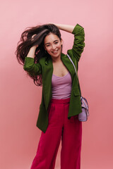 Playful asian young woman posing on pink background holding dark fluttering hair in her hands. In trendy bright outfit of green jacket and crimson pants, beauty smiles sincerely.