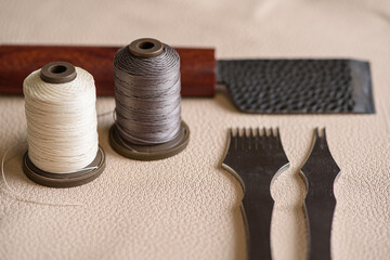 Fototapeta na wymiar Shoemaker tools for sewing handmade shoes in workshop. Punches, spools of thread and craft knife laying on beige genuine leather. Selective focus. Copy space.