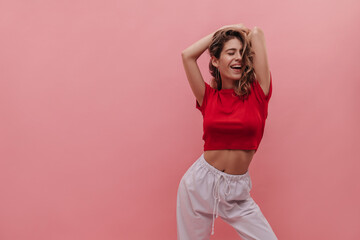 Slender young caucasian girl with curly hair is having fun on pink background. Pretty woman in red crop top with bare belly and white pants posing in studio. Active lifestyle concept