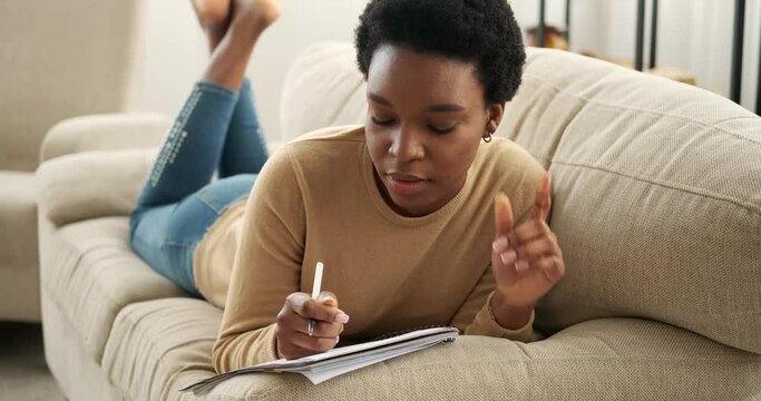 Thoughtful woman lying on sofa and writing in notepad at home