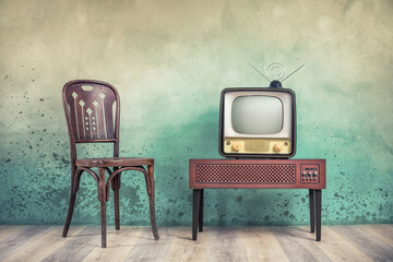 Television set receiver from 50s on wooden outdated analog TV stand and old classic chair front color textured aged concrete wall background. Vintage style filtered photo