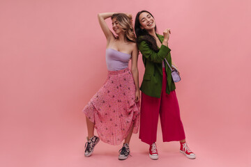 Full body shot of young european dancers girls posing in bright clothes against pink background. Gorgeous woman with wavy blonde hair and her brunette friend have fun in front of camera.