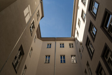 Fototapeta na wymiar Looking up between tall buildings to the blue sky. Facades of houses in front of the viewer. Sunlight is hitting the wall and windows are reflecting the light. A dark place in the backyard.