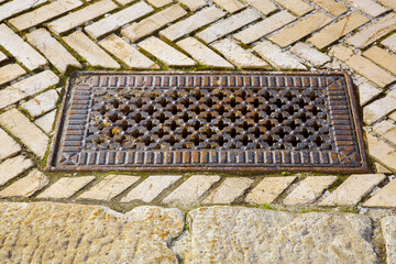 iron rusty rectangular manhole with shaped holes in form of crosses in lid on the sidewalk paved...