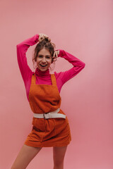 Vivid picture of estonian young woman playing with bun of her hair and smiling broadly against pink background. In vintage orange jumpsuit with pink long sleeves, blonde is looking at camera.