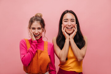 Beautiful young asian and european woman smile shyly with teeth on pink background. Portrait of two best friends in fashionable clothes showing joy and enjoying moments.