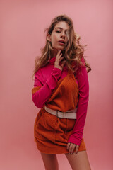 Fototapeta premium Young ukrainian lady posing alone over isolated pink background, thinking about date. Bright outfit of apricot denim dress and sweater, blonde loose curly hair with natural makeup.