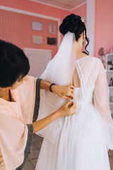 The bridesmaid helps in the morning, buttoning a button on the back of the bride on a white corset of a lace dress. Wedding portrait.