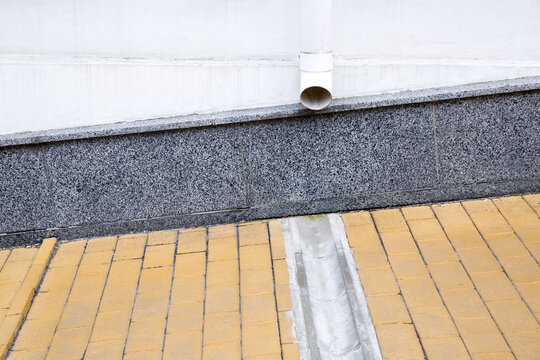 gray granite cladding on facade of a house wall with a drainage pipe and a concrete dish canal on a stone slab sidewalk to drain rainwater front view.