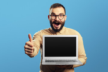 Young bearded man holding laptop and showing blank screen while showing thumb up wearing eyeglasses and beige shirt, isolated over blue wall