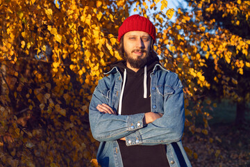 hipster man with a beard in jeans and knitted red hat is stand on a woodpile in autumn