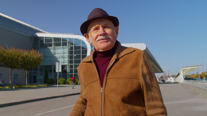 Portrait of senior pensioner tourist grandfather businessman stay near international airport hall or railway station carrying luggage suitcase waiting for plane or train. Travel, vacation, holidays