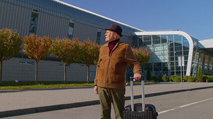 Portrait of elderly retired man tourist near airport terminal waiting boarding on plane for traveling. Stylish senior mature grandfather carrying luggage suitcases bag to railway station. Vacation