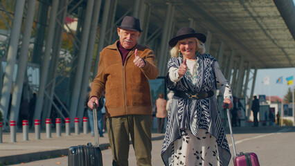 Senior happy pensioner tourists grandmother grandfather with luggage near international airport hall raises thumbs up agrees something or gives positive reply recommends advertisement likes good, ok