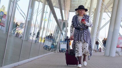 Senior tourist grandmother walking on international airport hall carrying luggage suitcase texting Messages on Smartphone and smiles. Travel, vacation. Pensioner business woman using mobile phone