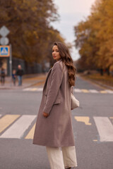 Elegant brunette woman with long wavy hair wearing grey wool coat, holding white handbag and walking at city street on autumn day. Portrait against yellow leaves