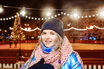 Happy smiling young Caucasian woman in scarf, hat, jacket, mittens by the outdoor rink. New year, fun, winter concept 