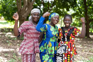Foto op Canvas Group of young black African villagers in colourful traditional dresses smiling at the camera with their clenched fists as a symbol for women's strength and gender equality © Riccardo Niels Mayer