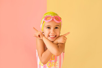 Cute adorable funny girl in a trendy Swim goggles and a Swimming Cap, pointing fingers in different sides at copy space on a pink and yellow studio background. Child indicating at discount or offer