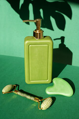 Rectangular green bottle with a jade roller massager and gua sha scraper for face massage on a...
