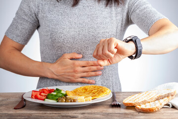 Intermittent fasting concept with a woman sitting hungry in front of food and looking at her watch...