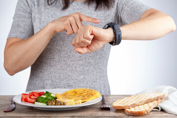 Intermittent fasting concept with a woman sitting hungry in front of food and looking at her watch to make sure she breaks fast on the correct time. A dietary modification for healthy lifestyle. - obrazy, fototapety, plakaty