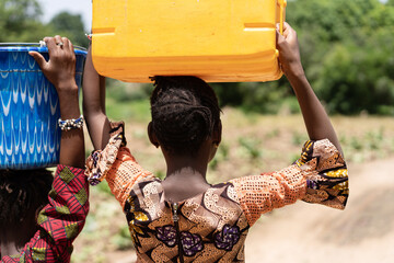 Rear view of a young black African girl in a colouful dress with a huge water can on her head walking on dirt road