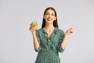 Beautiful young woman with fresh lemonade showing victory gesture on grey background