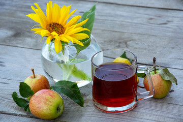 Still life with a glass of tea, sunflower flowers in a vase and apples on a wooden background.