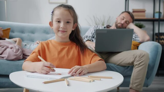 Slow motion portrait of cute little girl smiling drawing looking at camera while parents are sleeping in background. People and family life concept.