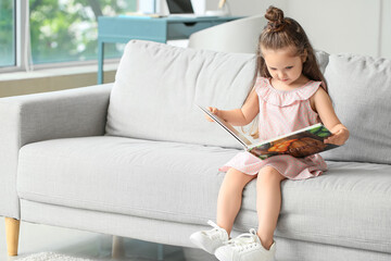 Adorable little girl reading book on sofa at home