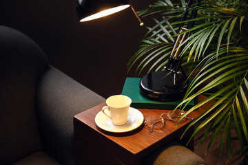 Stand with cup, eyeglasses and glowing lamp on sofa in dark room
